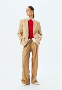 Beige oversized blazer, rode ribbed trui, witte button-down shirt en tan wijde broek. Inclusief gouden oorbellen en witte schoenen.