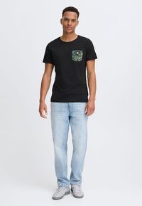 Camiseta negra de manga corta con un bolsillo estampado en verde y azul, combinada con jeans de un azul claro y zapatillas grises.