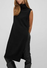 Svart ärmlös polokragnklänning med ribbade detaljer och sidoklyfta, tillverkad av ett mjukt stickat material, faller till mitten av låret. Kombineras med svarta läderbyxor.