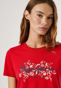 T-shirt rouge à manches courtes avec un col rond, présentant un motif floral blanc et un texte bleu indiquant "Pepe Jeans LONDON". En coton.