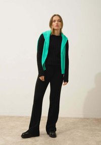 Pull en maille côtelée noir et pantalon assorti, superposé avec un gilet en polaire turquoise vif. Des chaussures noires massives complètent la tenue.