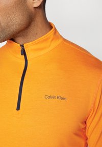 Oranžni pulover z zadrgo na vratu, ki ima mornarsko modro zadrgo in vezeno oznako "Calvin Klein" na prsih, narejen iz gladkega, teksturiranega materiala.