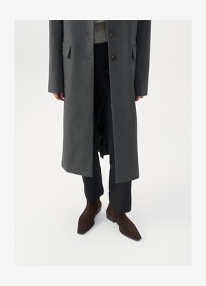 Gabardina gris larga con dos botones, ajuste entallado, bolsillos laterales; llevada sobre un suéter gris y pantalones negros, combinada con botines de ante marrón.