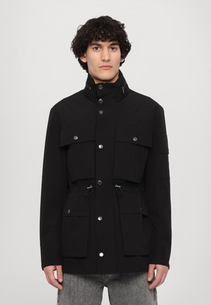 Black utility jacket featuring a high collar, multiple front pockets, adjustable waist with drawstrings, and snap button accents.