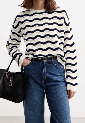 Kobieta ubrana w kremowy i granatowy sweter w faliste paski, niebieskie dżinsy z czarnym paskiem, trzymająca czarną torebkę w jednej ręce.