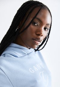 Ανοιχτό μπλε hoodie με κορδόνι, με λευκό σχέδιο κειμένου και μαλακή υφή υφασμάτων, τονίζοντας μια κοντινή λήψη χαρακτηριστικών προσώπου και πλεγμένων μαλλιών.