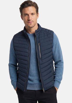 Mann trägt eine marineblaue gesteppte Weste über einem hellblauen Strickpullover, die Hände in den Taschen einer schwarzen Hose, vor einem schlichten weißen Hintergrund.