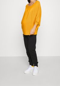 Personne enceinte portant un pull ample jaune moutarde, pantalon noir, baskets blanches, debout devant un fond clair uni.