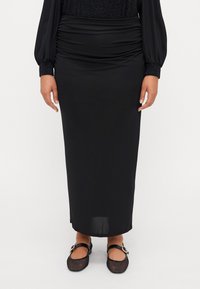 Svart, knelang skjørt laget av glat stoff med rynket detaljering i midjen; parret med svarte mesh ballerinaer med spenne.