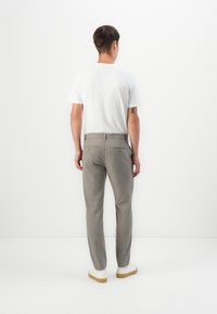 Hombre de espaldas con camiseta blanca, pantalones grises de corte recto, zapatillas blancas y un tatuaje visible en su brazo izquierdo, de pie frente a un fondo blanco.