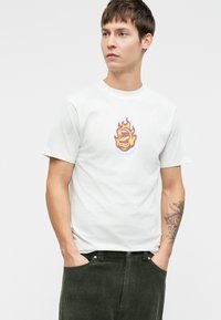 Tricou alb din bumbac cu un grafic colorat de flacără, având o gură și textul 'Santa Cruz', asortat cu pantaloni din corduroy verde închis.