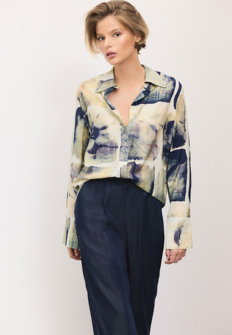 Multi-colored, long-sleeve shirt with an abstract print, button-down collar, and soft fabric. Paired with dark, loose-fitting trousers.