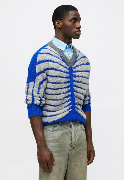 Μπλε και γκρι ριγέ πουλόβερ με V-λαιμό, υφή πλεκτού, ριμπ μανσέτες, φορεμένο πάνω από μια ανοιχτό μπλέ πουκαμίσα, συνδυασμένο με ξεθωριασμένα τζιν.