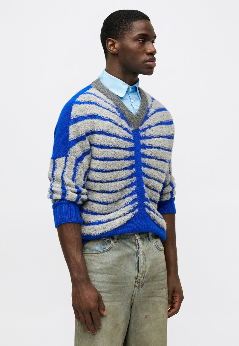 Suéter a rayas azul y gris con escote en V, patrón de tejido texturizado, puños de canalé, llevado sobre una camisa azul clara, combinado con jeans desgastados.