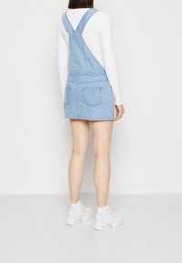 Denim overall dress in light blue with adjustable straps, a fitted waist, and a mini length, featuring two back pockets and a soft texture.