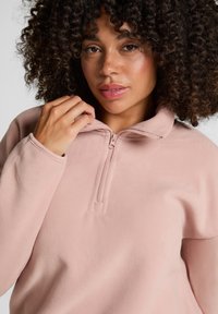 Jasnoróżowy polarowy pulower na zamek błyskawiczny z wysokim kołnierzem, z półdługościoym zamkiem i miękką strukturą. Widoczne detale ściegów na kołnierzu.