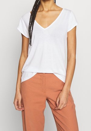 Kobieta z zaplecionymi włosami, ubrana w białą koszulkę z dekoltem w kształcie litery V, wpuszczoną w wysokie, rdzawe spodnie, stojąca na tle jednolitego tła.