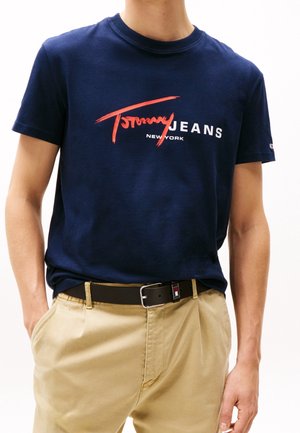 Homme portant un t-shirt Tommy Jeans bleu marine avec un logo rouge et blanc, un pantalon beige et une ceinture noire avec boucle argentée.