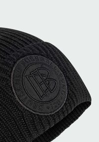 Sort strikket beanie med et tekstureret mønster og en cirkulær sort lap med præget logo, der tilføjer detaljer til det overordnede design.