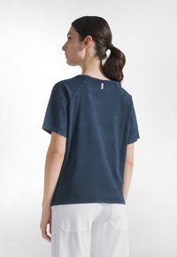 Kobieta o ciemnych włosach związanych w kucyk, ubrana w granatową koszulkę z krótkim rękawem i białe spodnie, stojąca tyłem na jednolitym jasnym tle.