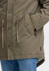 Parka vert olive avec une poche zippée, des cordons de serrage et des accents marron. Présente des boutons métalliques et un ourlet légèrement arrondi.