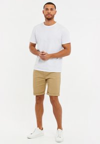 Camiseta blanca de cuello redondo combinada con pantalones cortos beige con dobladillos enrollados. El conjunto se completa con zapatillas blancas. Diseño sencillo y casual.