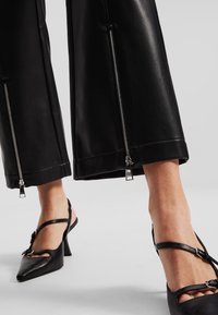 Zwarte leren broek met brede pijpen en ritssluitingen aan de onderkant, met een gladde textuur. Gecombineerd met zwarte sandaaltjes met bandjes.