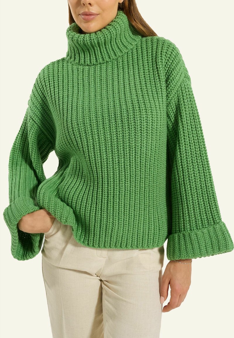 Frau trägt einen voluminösen grün gestrickten Rollkragenpullover mit weiten Ärmeln und hellbeigen Hosen, eine Hand in der Tasche.