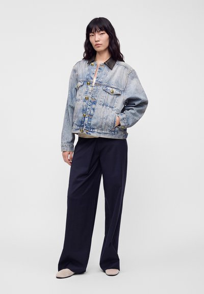 Femme portant une veste en jean bleu clair avec des boutons dorés, un pantalon noir à jambes larges et des chaussures beige, debout avec une main dans la poche.