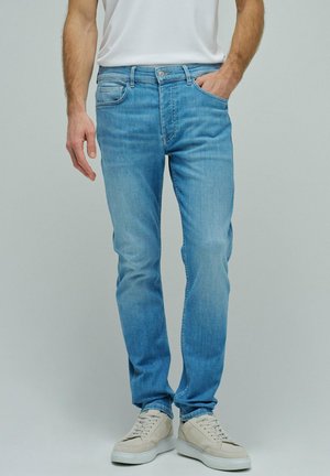 Hombre con vaqueros ajustados azul claro con la mano en el bolsillo y zapatillas blanco roto, de pie frente a un fondo liso gris claro.