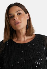 Svart paljetttopp med vertikal randdesign, med korta genomskinliga ärmar och rund halsringning. Bärs med långa svarta pärlörhängen.