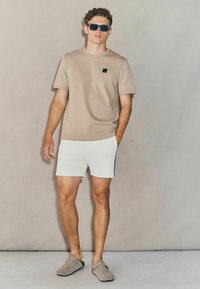 Camiseta de manga corta beige con un pequeño logotipo, pantalones cortos deportivos blancos y zapatos slip-on de ante gris. Modelo con gafas de sol negras.