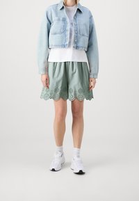 Personne portant une veste en denim bleu clair cropped, un t-shirt blanc, un short vert à ourlet festonné avec un motif découpé, des chaussettes blanches et des baskets.