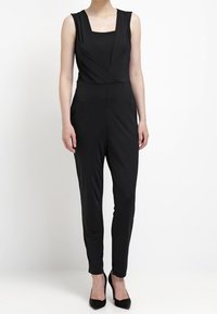 Combinaison noire avec un décolleté carré, sans manches, taille ajustée et jambes fuselées. Texture lisse avec un look minimaliste.