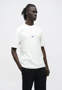 Νέος άνδρας με dreadlocks που φοράει λευκό κοντομάνικο πουκάμισο με μικρό μπλε λογότυπο και μαύρο παντελόνι, στέκεται με τα χέρια στις τσέπες.