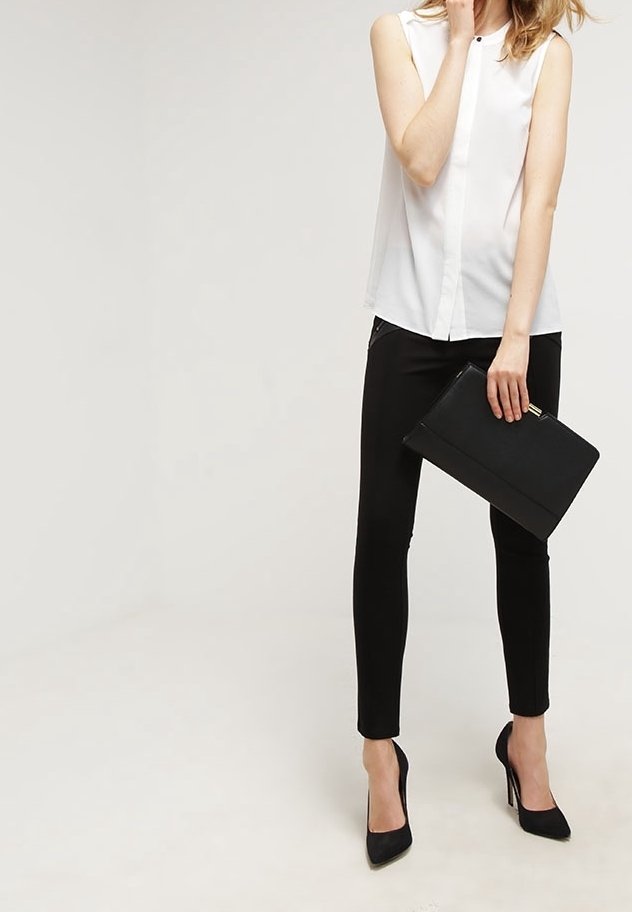 Vit utan ärm-blus med knäppning framtill, kombinerad med svarta tighta byxor och svarta högklackade skor, som håller en svart kuvertväska.