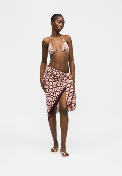 Γυναίκα που φοράει μπικίνι με καφέ και λευκό μοτίβο και αντίστοιχο σερφόν με σκίσιμο, στέκεται ξυπόλητη σε απλό ανοιχτό φόντο.