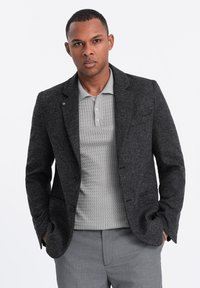 Blazer en laine gris foncé à deux boutons, porté sur un polo texturé gris clair. Le pantalon est à motifs gris clair avec une coupe ajustée.