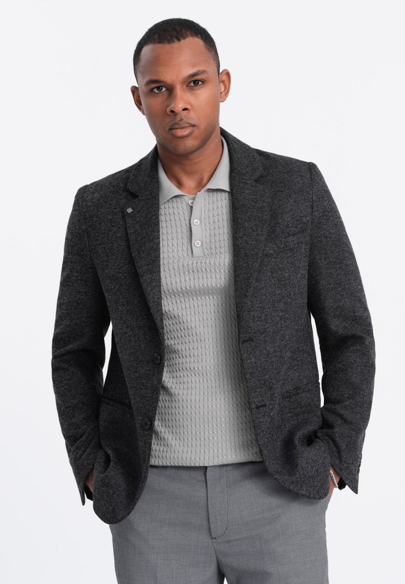 Blazer en laine gris foncé à deux boutons, porté sur un polo texturé gris clair. Le pantalon est à motifs gris clair avec une coupe ajustée.