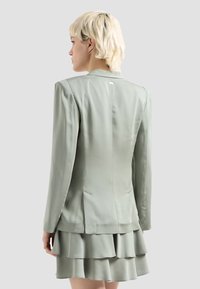 Blazer in satin verde chiaro con vestibilità sartoriale, maniche lunghe e un'unica spacco posteriore. Presenta un logo discreto vicino allo scollo.