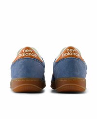 Blue suede sneakers with orange accents on the heel, featuring a rounded toe and a textured rubber sole. “New Balance” logo displayed prominently.