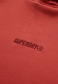 Sudadera roja con una textura suave que presenta un logo en relieve de "SUPERDRY". La tela parece lisa y está diseñada para un uso casual.