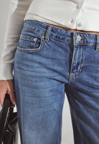 Suorat siniset farkut, joissa on kaksi etutaskua, metallinen nappisuljenta ja hillitty kulutustyyli. Yhdistetty valkoiseen paitaan.