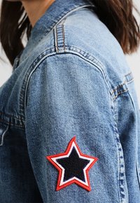 Jeansjacka med medelblå tvätt, med ett röd stjärn-patch omgiven av vitt och svart på vänster ärm, med synlig sömnad.