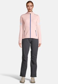 Giacca zip-up rosa chiaro con dettagli bianchi e blu, caratterizzata da un collo alto e un design aderente. Abbinata a pantaloni grigi scuri e stivali chiari.