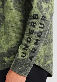 Zelená maskáčová košile s dlouhým rukávem a texturovanou tkaninou, která má černé logo "UNDER ARMOUR" potisknuté vertikálně na rukávu.