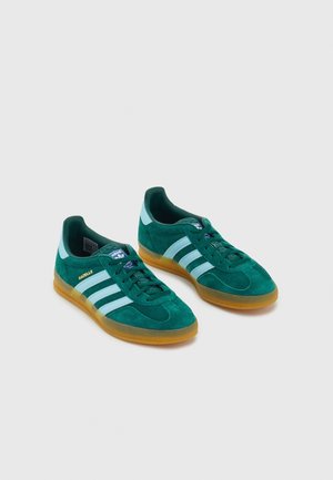 Par grønne Adidas Gazelle sneakers med hvide striber og såler af gummi på en ensfarvet lys baggrund.