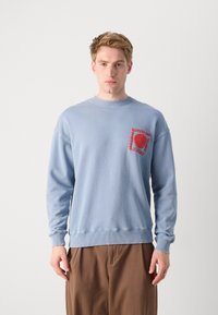 Light blue crewneck sweatshirt made of cotton, featuring a red graphic design on the chest pocket area and ribbed cuffs and hem.