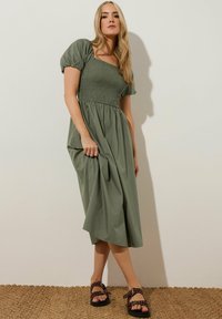 Olivegrünes, knielanges Kleid mit gerafftem Oberteil, Puffärmeln und fließendem Rock. Kombiniert mit braunen, mit Nieten besetzten Sandalen.