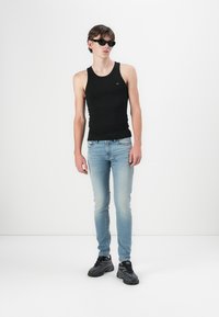 Top negru fără mâneci, blugi slim-fit de culoare albastră deschisă și pantofi sport negri. Model purtând ochelari de soare întunecați; ținută simplă și casual pe un fundal neutru.